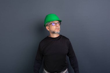 Resme poz veren yeşil kasklı ve siyah elbiseli bir memur portresi. Gri arka planda izole. İnşaat işçisi.