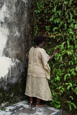 Köle gibi giyinmiş, zincirlenmiş ve ağzı ve boynunda demir bir maskeyle yeşil bitkilerle eski bir duvara yaslanmış bir kadın. Brezilya 'da kölelik. Köle Anastacia 'nın temsilcisi.