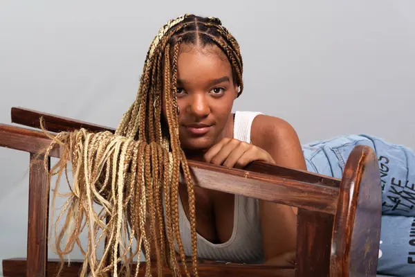 Saçında örgüleri olan güzel esmer bir kadın stüdyoda oturmuş, ahşap bir tabureye yaslanmış kameraya bakıyor. Sakin ve kendinden emin biri. Gri arkaplanda izole.