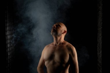 Olgun, üstsüz, kel ve sakallı bir adam fotoğrafı için poz veriyor. Siyah arka planda dumanla izole edilmiş. Güney Amerika