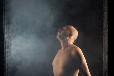 Olgun, üstsüz, kel ve sakallı bir adam fotoğrafı için poz veriyor. Siyah arka planda dumanla izole edilmiş. Güney Amerika