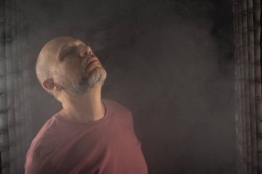 Kel ve sakallı bir adam stüdyoda siyah arka planda duman içinde hareketler ve ifadeler yapıyor. Güney Amerika