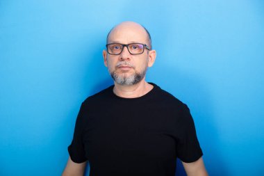 Sakallı, kel, reçeteli gözlüklü ve siyah gömlekli bir adam mavi arka planda fotoğraf için poz veriyor. Stüdyo çekimi.