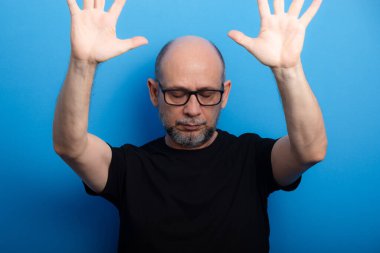 Sakallı, kel, reçeteli gözlük takan ve kolları kameraya dönük siyah gömlekli bir adam. Kendinden emin ve sakin. Stüdyo çekimi.