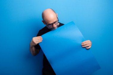 Sakallı, kel, reçeteli gözlüklü, siyah gömlekli, elinde mavi bir afişe bakan bir adam. Kendinden emin ve odaklanmış. Stüdyo çekimi.