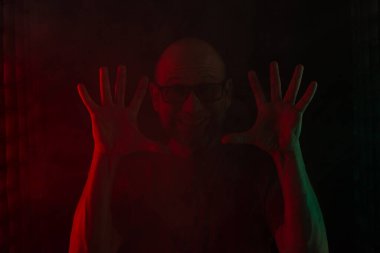 Reçeteli gözlüklü kel ve sakallı bir adam kameraya gülümsüyor ve iki elini de gösteriyor. Koyu kırmızı arka planda izole edilmiş. Güney Amerikalı..