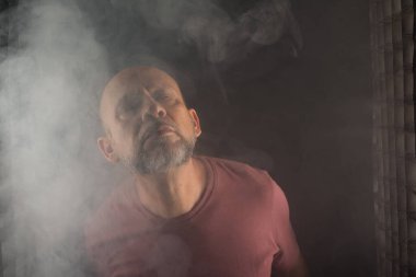 Kel ve sakallı bir adam stüdyoda siyah arka planda duman içinde hareketler ve ifadeler yapıyor. Güney Amerika