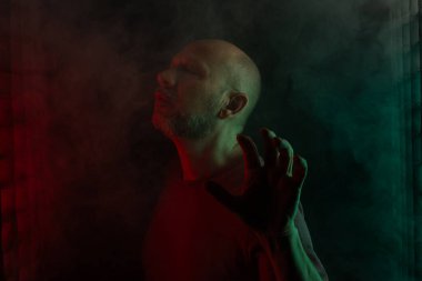 Beyaz, kel ve sakallı adam portresi, kameraya yüz ifadeleri yapıyor. Siyah dumanlı arka planda, renkli ışıklarla izole edilmiş. Güney Amerikalı..