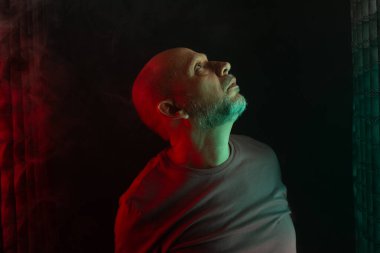 Kel ve sakallı adam yukarı bakıyor. Koyu dumanlı arka planda, renkli ışıklarla izole edilmiş. Güney Amerikalı..