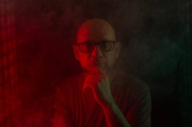 Gözlüklü kel ve sakallı bir adam kameraya bakıyor. Koyu dumanlı arka planda, renkli ışıklarla izole edilmiş. Güney Amerikalı..