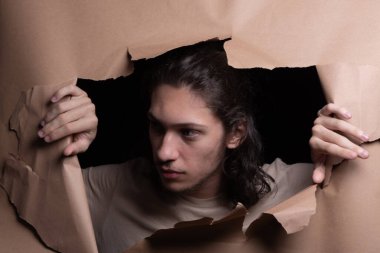 Man's head sticking out and looking through a paper hole with his hands pushing the paper aside. Studio portrait. clipart