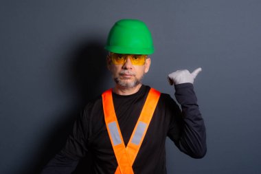 Yeşil kask, yansıtıcı yelek ve koruyucu gözlük takan bir memur parmak hareketi yapıyor. Gri arka planda izole. İnşaat işçisi.