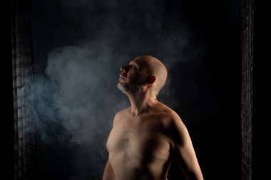 Olgun, üstsüz, kel ve sakallı bir adam fotoğrafı için poz veriyor. Siyah arka planda dumanla izole edilmiş. Güney Amerika