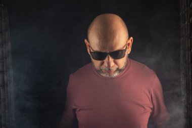 Güneş gözlüğü takan kel ve sakallı bir adam. Stüdyoda siyah dumanlı bir arka planı olan bir fotoğraf için poz veriyor. Güney Amerika