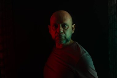 Beyaz, kel ve sakallı adam kameraya bakıyor. Siyah arka planı ve renkli ışıkları olan bir stüdyo portresi. Güney Amerikalı..