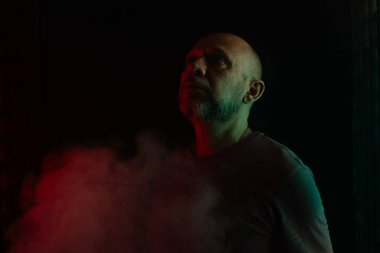 Beyaz, kel ve sakallı adam portresi, kameraya yüz ifadeleri yapıyor. Siyah dumanlı arka planda, renkli ışıklarla izole edilmiş. Güney Amerikalı..