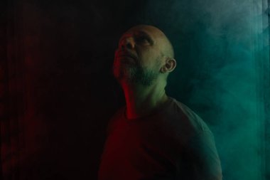 Kel ve sakallı adam yukarı bakıyor. Koyu dumanlı arka planda, renkli ışıklarla izole edilmiş. Güney Amerikalı..