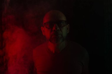 Gözlüklü kel ve sakallı bir adam kameraya bakıyor. Koyu dumanlı arka planda, renkli ışıklarla izole edilmiş. Güney Amerikalı..