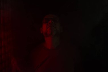 Kel ve sakallı bir adam, reçeteli gözlüklü, koyu kırmızı bir geçmişi olan yoğun duman arasında fotoğraf için poz veriyor. Güney Amerikalı..