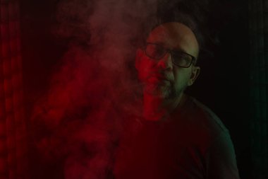 Gözlüklü kel ve sakallı bir adam kameraya bakıyor. Koyu dumanlı arka planda, renkli ışıklarla izole edilmiş. Güney Amerikalı..