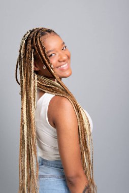 Saçları örgülü, kot pantolon ve beyaz gömlek giyen, kameraya bakıp gülümseyen güzel esmer kadın. Gri stüdyo arka planında izole edilmiş..