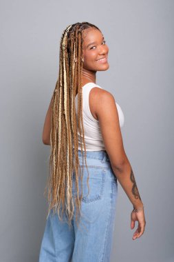 Saçları örgülü, kot pantolon ve beyaz gömlek giyen, kameraya bakıp gülümseyen güzel esmer kadın. Gri stüdyo arka planında izole edilmiş..