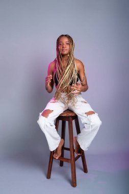 Saçında örgüler, beyaz kot pantolon ve siyah bluz giyen esmer bir manken, tahta bir taburede kameraya poz veriyor. Gri bir arkaplanda izole edilmiş.