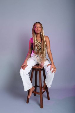 Saçında örgüler, beyaz kot pantolon ve siyah bluz giyen esmer bir manken, tahta bir taburede kameraya poz veriyor. Gri bir arkaplanda izole edilmiş.