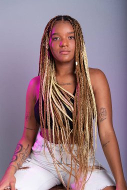 Saçında örgüler, beyaz kot pantolon ve siyah bluz giyen esmer bir manken, tahta bir taburede kameraya poz veriyor. Gri bir arkaplanda izole edilmiş.