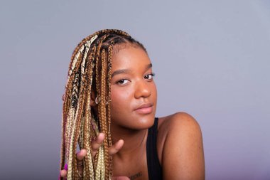 Saçında örgüler olan esmer bir model, siyah bir bluz giyiyor ve kameraya bakıyor. Kendine güvenen ve sakin biri. Gri arkaplanda izole.