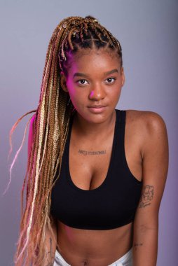 Saçında örgüler olan esmer bir model, siyah bir bluz giyiyor ve kameraya bakıyor. Kendine güvenen ve sakin biri. Gri arkaplanda izole.