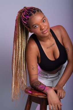 Saçında saç örgüleri olan esmer bir model, beyaz kot pantolon ve kameraya poz veren tahta bir tabureye yaslanmış siyah bir bluz giyiyor. Gri bir arkaplanda izole edilmiş.