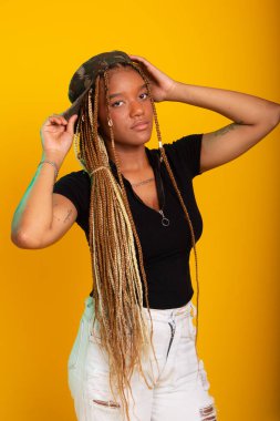 Saçında örgüleri olan, üzerinde pantolon ve bluzla kameraya poz veren güzel bir genç kadının stüdyo portresi. Sarı arkaplanda izole edilmiş.