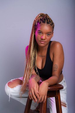 Saçında saç örgüleri olan esmer bir model, beyaz kot pantolon ve kameraya poz veren tahta bir tabureye yaslanmış siyah bir bluz giyiyor. Gri bir arkaplanda izole edilmiş.