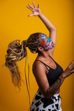 Renkli yüz boyası ve saçlarında örgülerle kameraya poz veren güzel bir genç kadın. Sarı arkaplanda izole edilmiş.