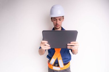 Genç mimar beyaz kask ve parlak yelek giyiyor, dizüstü bilgisayarla çalışıyor. Beyaz arka planda izole edilmiş. Güvenlik ekipmanları.