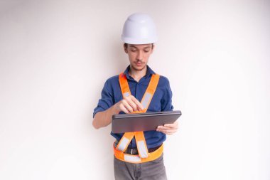 Genç mimar beyaz kask ve parlak yelek giyiyor, dizüstü bilgisayarla çalışıyor. Beyaz arka planda izole edilmiş. Güvenlik ekipmanları.