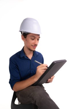 Genç mimar beyaz kask takıyor, oturuyor ve tabletle çalışıyor. Beyaz arka planda izole edilmiş. Güvenlik ekipmanları.