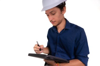 Genç mimar beyaz kask takıyor, oturuyor ve tabletle çalışıyor. Beyaz arka planda izole edilmiş. Güvenlik ekipmanları.