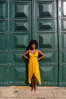 Sarı elbiseli, afro saçlı güzel siyah bir kadın büyük yeşil bir kapıya yaslanmış bir fotoğraf için poz veriyor. Mutlu ve gülümsüyor