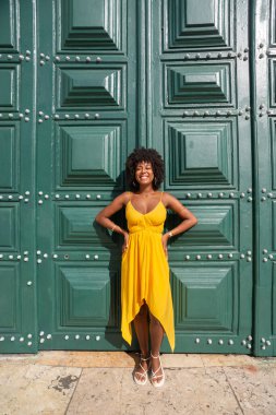 Sarı elbiseli, afro saçlı güzel siyah bir kadın büyük yeşil bir kapıya yaslanmış bir fotoğraf için poz veriyor. Mutlu ve gülümsüyor