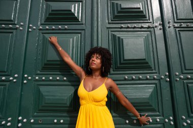 Sarı elbiseli, afro saçlı güzel siyah bir kadın büyük yeşil bir kapıya yaslanmış bir fotoğraf için poz veriyor. Mutlu ve gülümsüyor