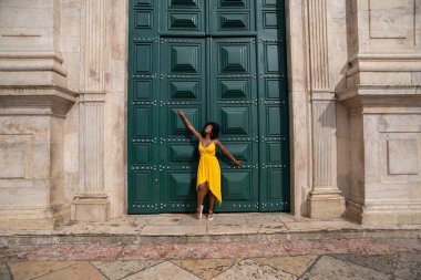 Sarı elbiseli, afro saçlı güzel siyah bir kadın büyük yeşil bir kapıya yaslanmış bir fotoğraf için poz veriyor. Mutlu ve gülümsüyor