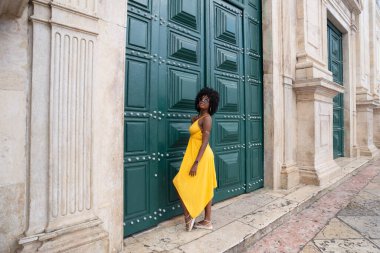 Sarı elbiseli, afro saçlı güzel siyah bir kadın büyük yeşil bir kapıya yaslanmış bir fotoğraf için poz veriyor. Mutlu ve gülümsüyor