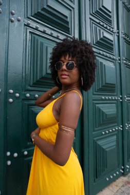 Siyah saçlı, sarı elbiseli ve güneş gözlüklü güzel bir kadın büyük yeşil bir kilise kapısının yanında fotoğraf için poz veriyor. Mutlu ve gülümsüyor