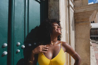 Siyah saçlı, sarı elbiseli güzel bir zenci kadın büyük yeşil kilise kapısına poz veriyor. Mutlu ve gülümsüyor