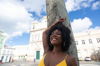 Afro saçlı, sarı elbiseli, fotoğraf için poz veren güzel siyah kadın kilise arka planında hindistan cevizi ağacı gövdesine yaslanıyor. Mutlu gülümseyen kişi