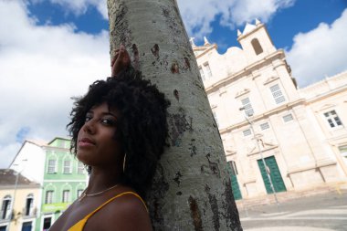 Afro saçlı, sarı elbiseli, fotoğraf için poz veren güzel siyah kadın kilise arka planında hindistan cevizi ağacı gövdesine yaslanıyor. Mutlu gülümseyen kişi