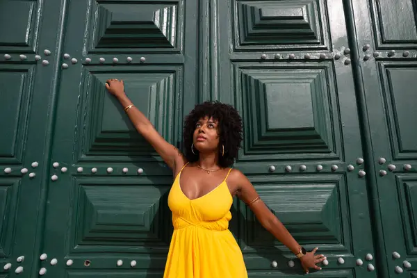 Sarı elbiseli, afro saçlı güzel siyah bir kadın büyük yeşil bir kapıya yaslanmış bir fotoğraf için poz veriyor. Mutlu ve gülümsüyor