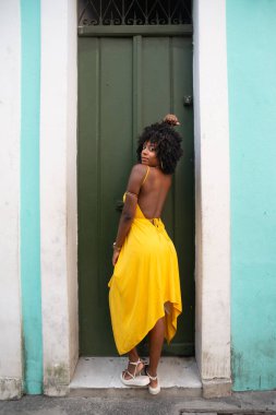 Afro saçlı, sarı elbiseli, yeşil kapıya dayanmış fotoğraf için poz veren güzel siyahi kadın portresi. Yolculukta mutlu insan.
